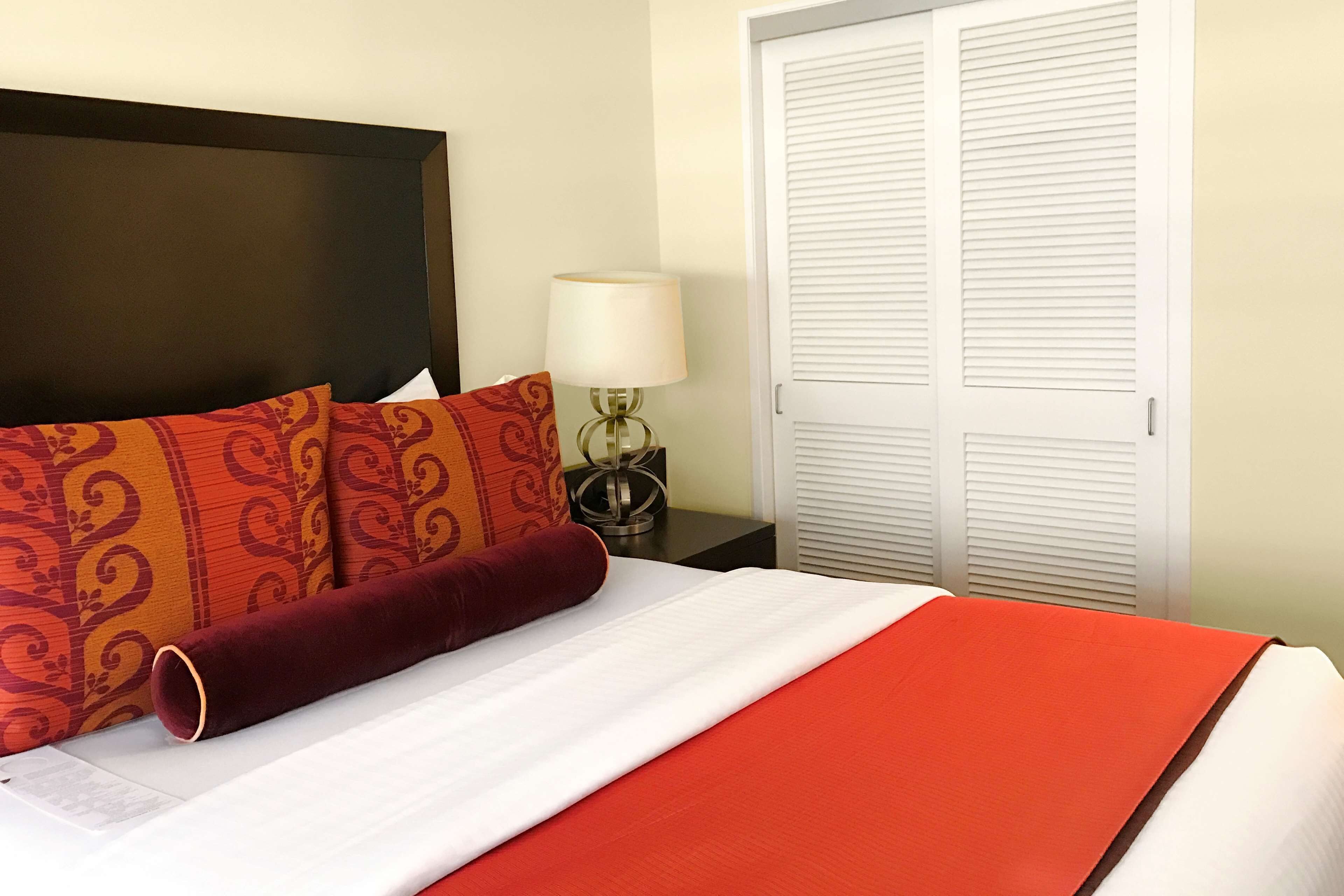 Una cama bien hecha con almohadas estampadas y un detalle rojo, una lámpara de noche y un armario blanco con puertas batientes.