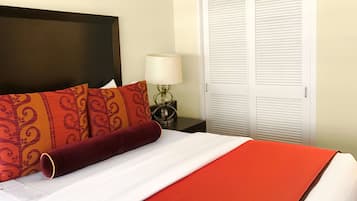 Una cama bien hecha con almohadas estampadas y un detalle rojo, una lámpara de noche y un armario blanco con puertas batientes.