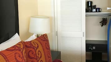 Una habitación de hotel con cama, mesita de noche con lámpara, armario y estante con cafetera y utensilios.