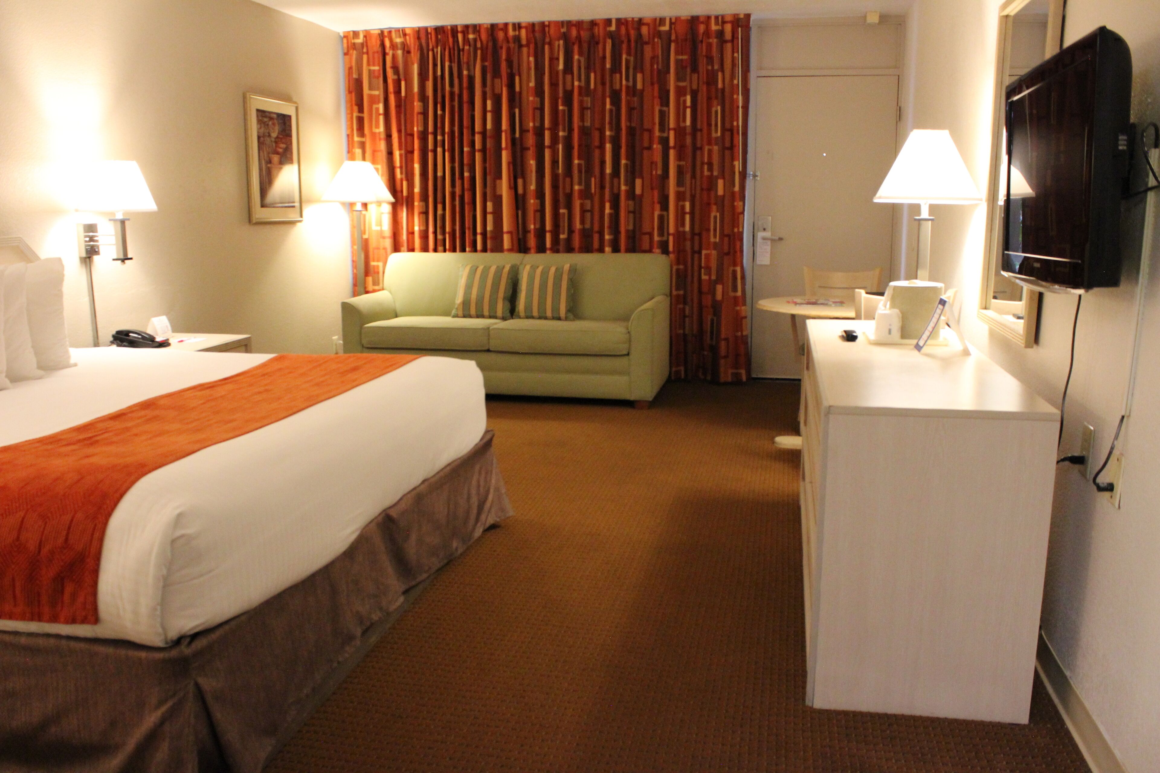 A hotel room with a bed, a green sofa, a desk with a lamp, and a television.