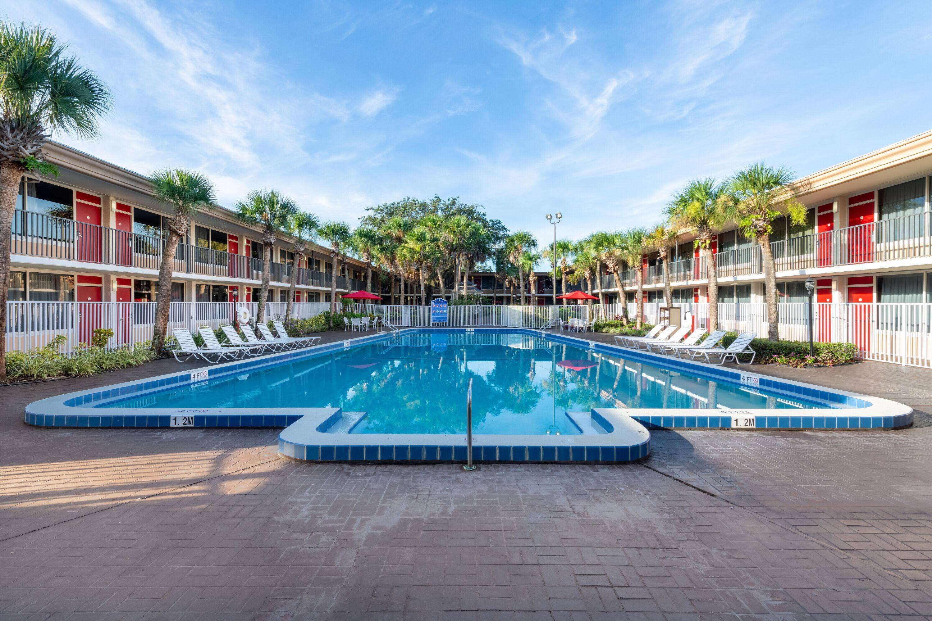 Une piscine entourée de chaises longues et de palmiers, elle-même bordée d’un hôtel aux murs extérieurs rouges et blancs.
