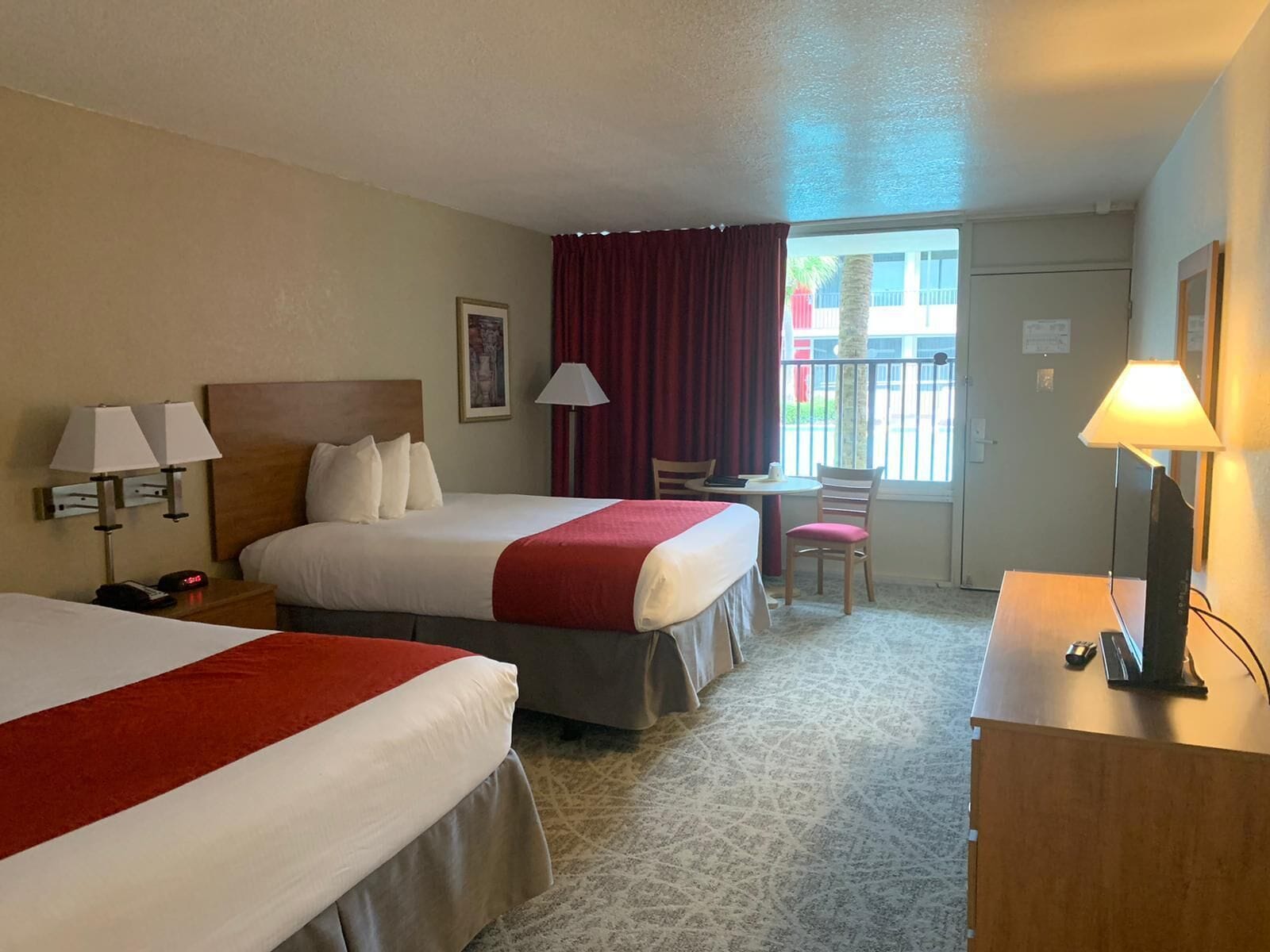 Ein Hotelzimmer mit zwei Betten, einem Schreibtisch, einem Stuhl, einer Lampe und einem Fenster mit Blick nach draußen.