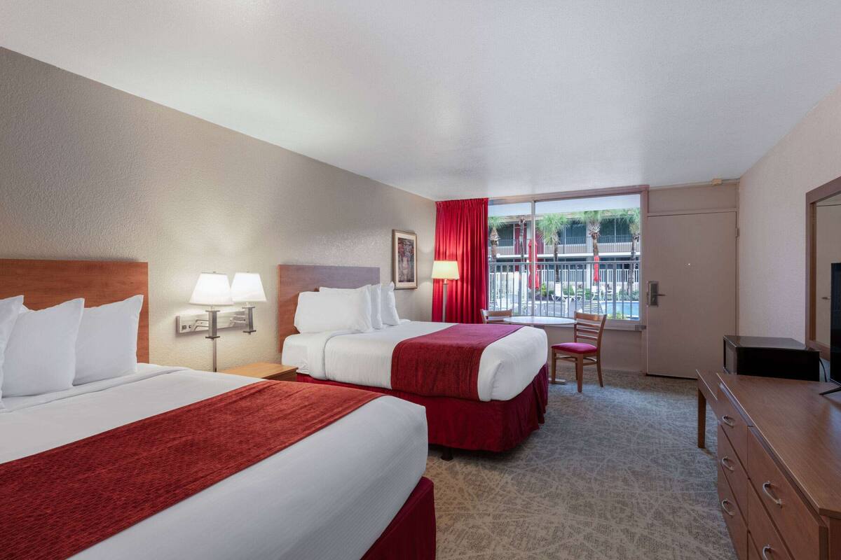 A hotel room with two beds, a desk, a chair, a TV, and a window with a view of palm trees and a balcony.