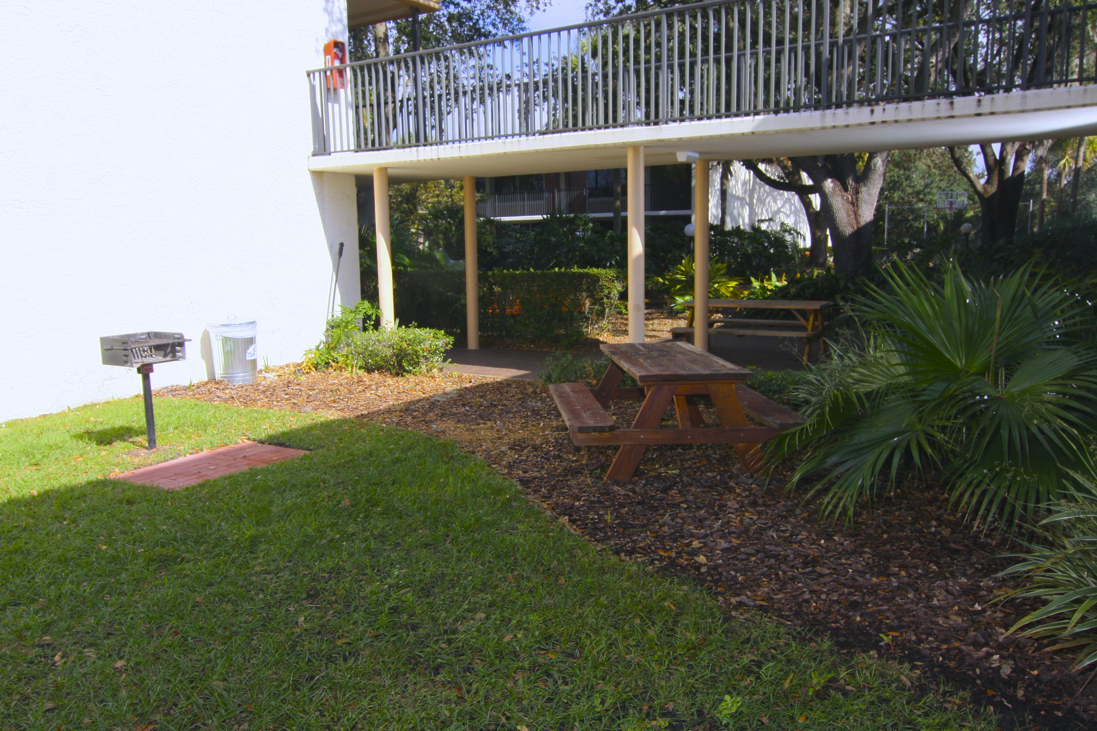 A covered outdoor area with picnic tables, a bench, and a white building with a balcony.