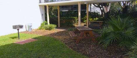 A covered outdoor area with picnic tables, a bench, and a white building with a balcony.