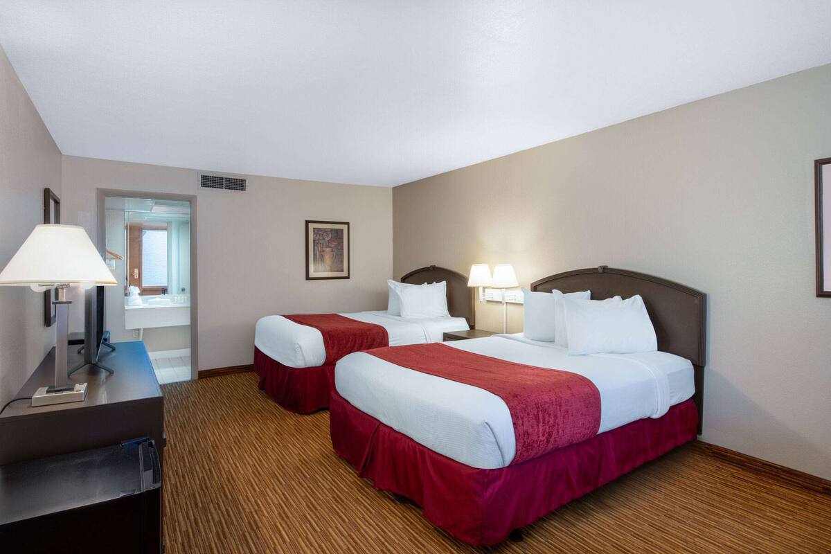 A hotel room with two beds, a desk with a lamp, and a view of the hallway.