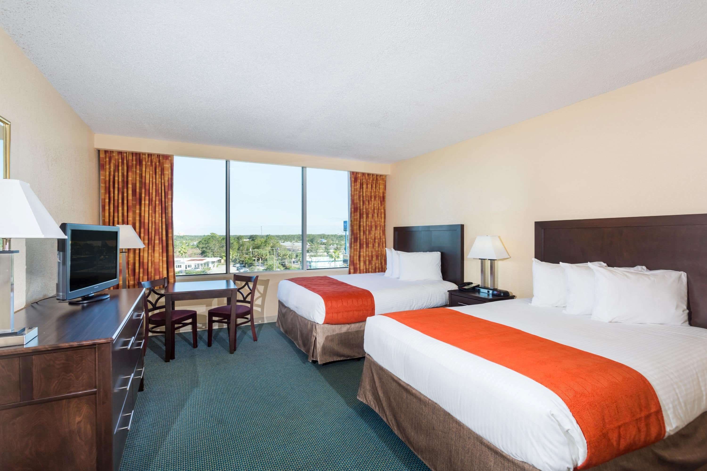A hotel room with two beds, a desk, a TV, and large windows.
