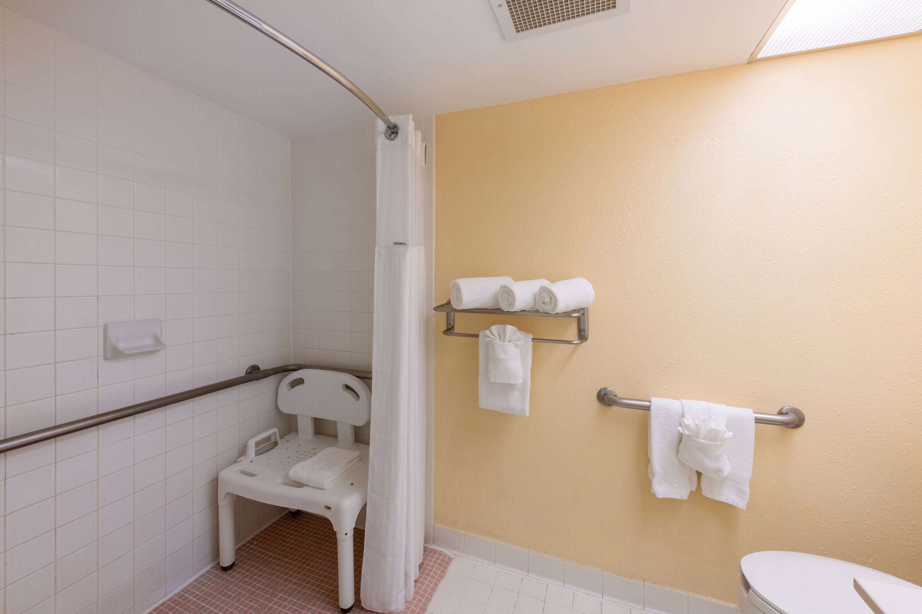 Een badkamer met een douche, toilet, een handgreep, een witte stoel en handdoeken op een rek.