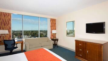 A hotel room with a large bed, a desk, a chair, a sofa, a TV, and a view of the city through large windows.