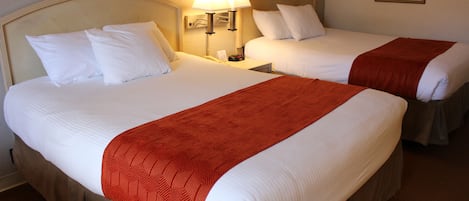 A hotel room with two beds, each with white bedding and a red throw blanket. There is a nightstand with a lamp between the beds.