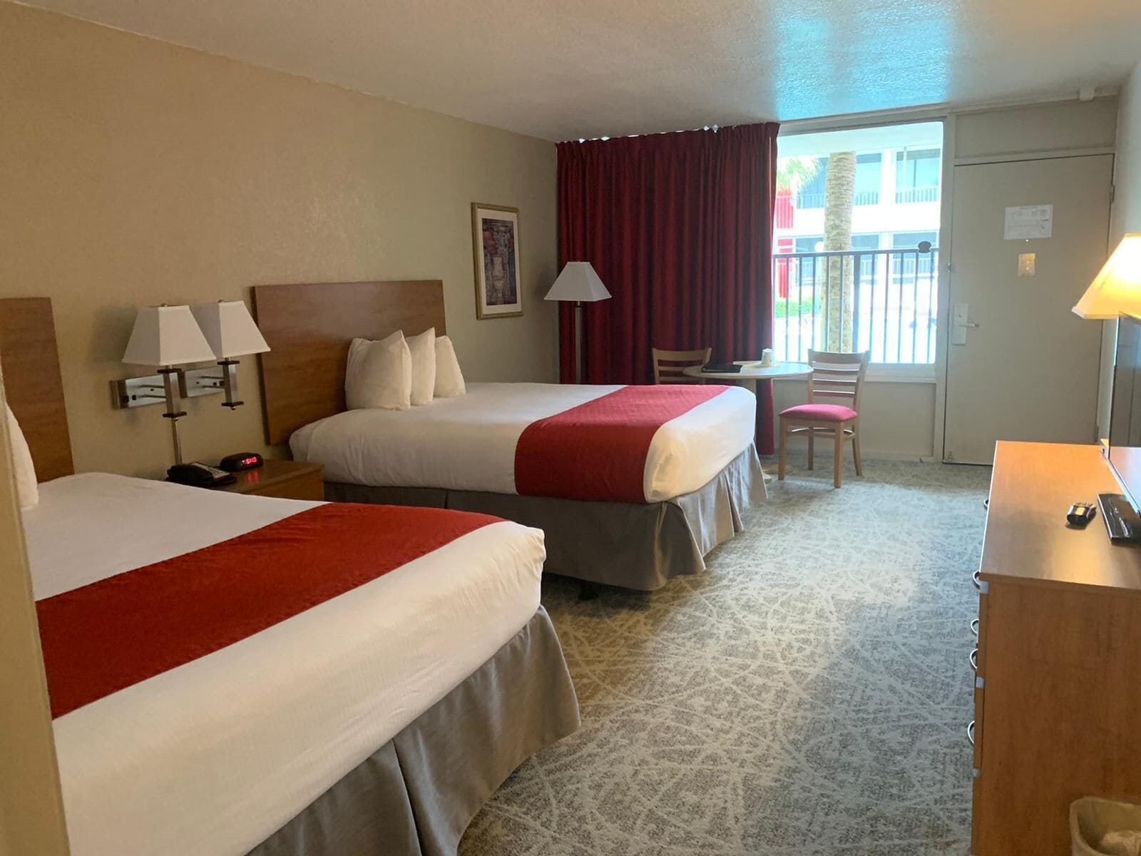 Une chambre d’hôtel avec deux lits, un bureau, une chaise, une lampe et une fenêtre avec des rideaux.