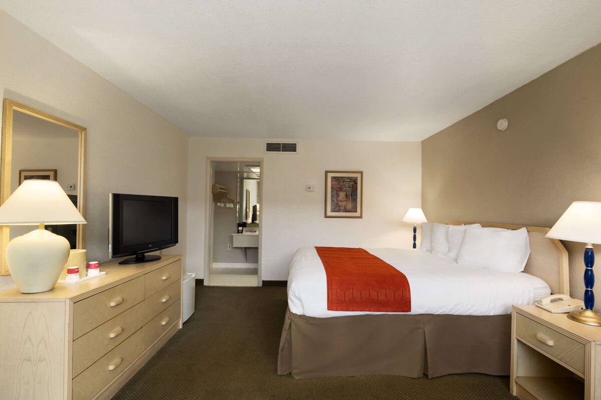 A hotel room with a bed, a television, a desk with a lamp, a dresser, and a mirror.