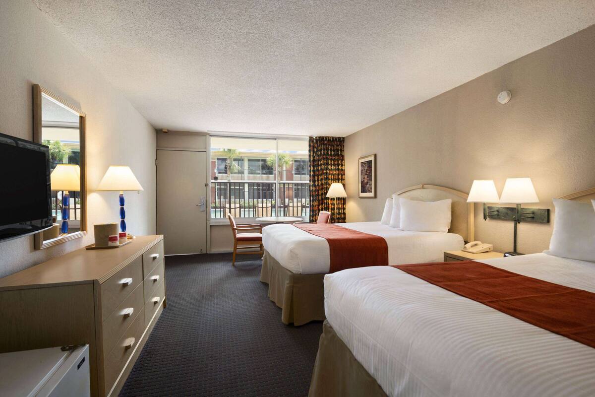 A hotel room with two beds, a desk, a television, and a window with a view of palm trees.