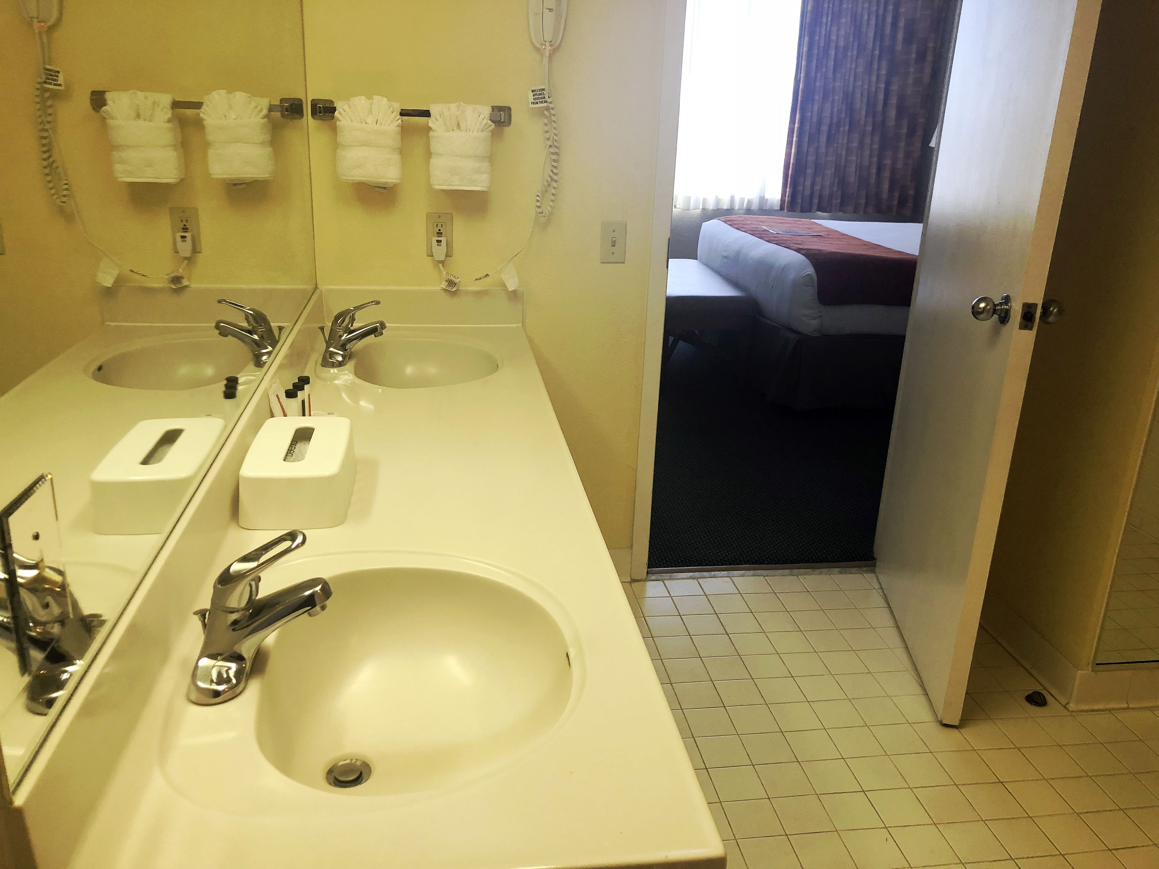 Een hotelbadkamer met twee wastafels, een spiegel en een bed dat door een open deur zichtbaar is.