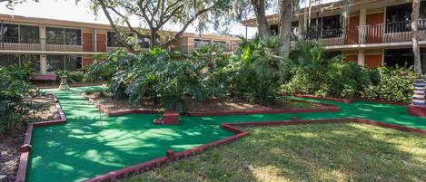 A miniature golf course with a green putting surface, surrounded by lush greenery and trees.