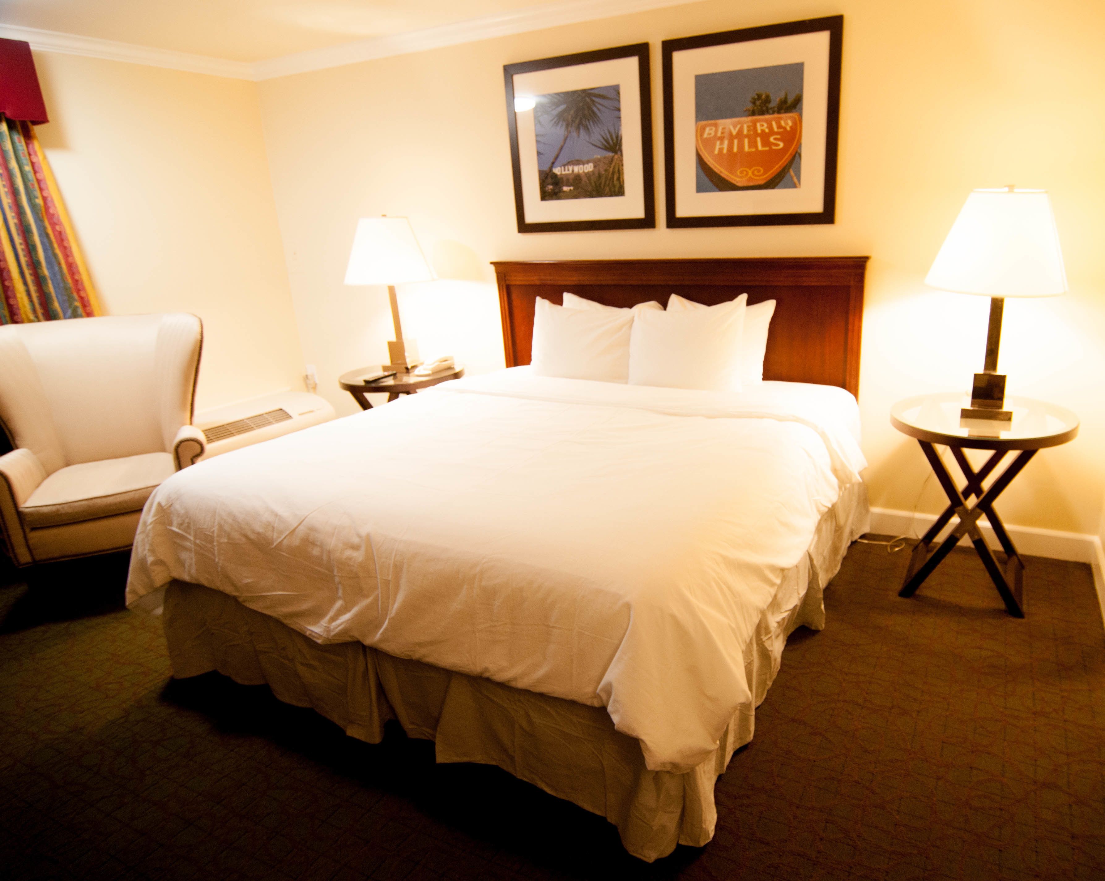 Habitación de hotel con una cama grande, una silla, dos mesitas de noche con lámparas y cuadros en la pared.