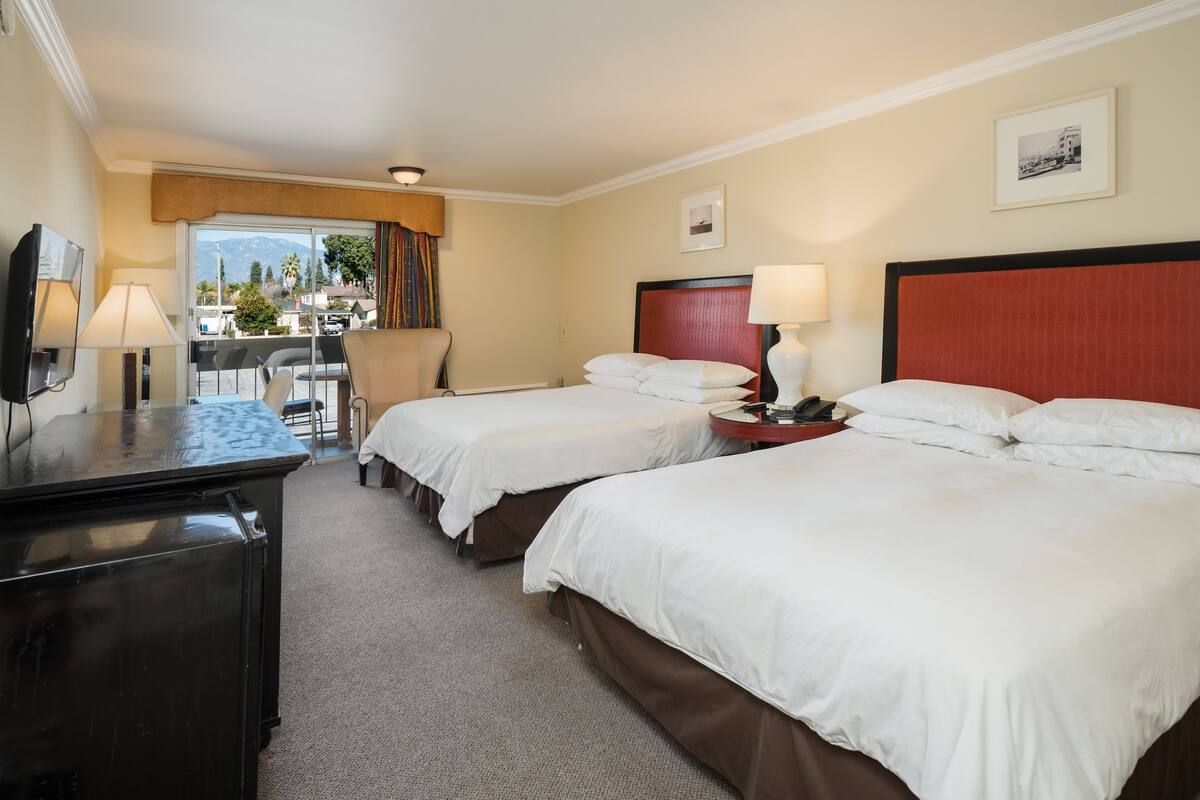 A hotel room with two beds, a desk, a chair, and a view of the outdoors.