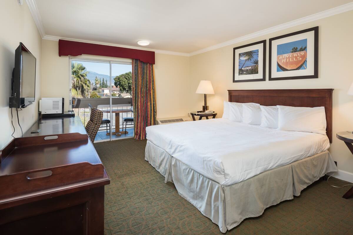 A hotel room with a large bed, a desk with a computer, a TV, and a balcony with a view of palm trees and mountains.
