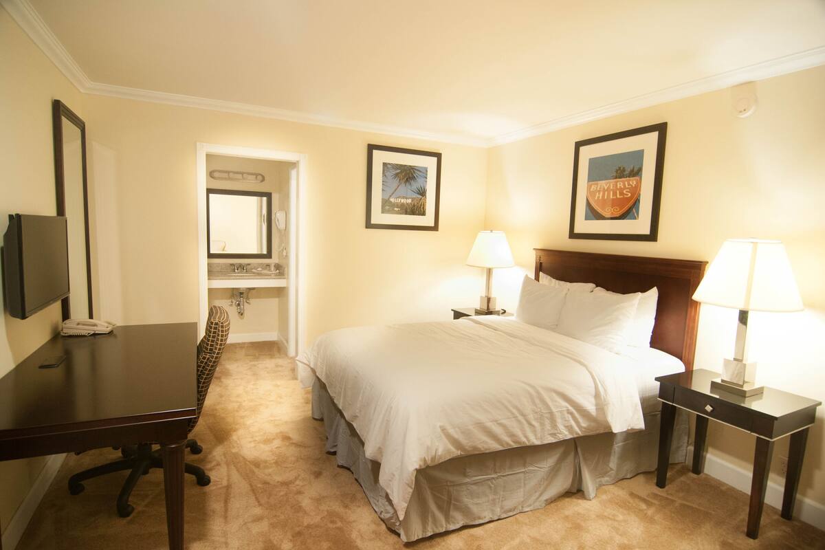 A hotel room with a bed, two bedside lamps, a desk with a chair, a television, and a framed picture on the wall.