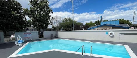 Una piscina rectangular con una solana con mosaicos, rodeada de un muro bajo y árboles.