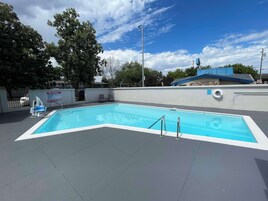 Una piscina rectangular con una solana con mosaicos, rodeada de un muro bajo y árboles.