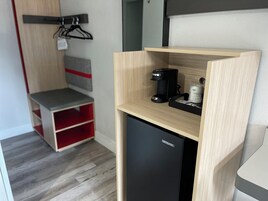 Una kitchenette compacta con cafetera, refrigerador pequeño y una estantería con bolsas de snacks.
