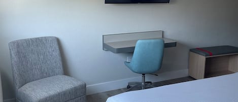 Quarto de hotel com uma cama, uma poltrona cinzenta, uma escrivaninha com cadeira e uma televisão montada na parede.