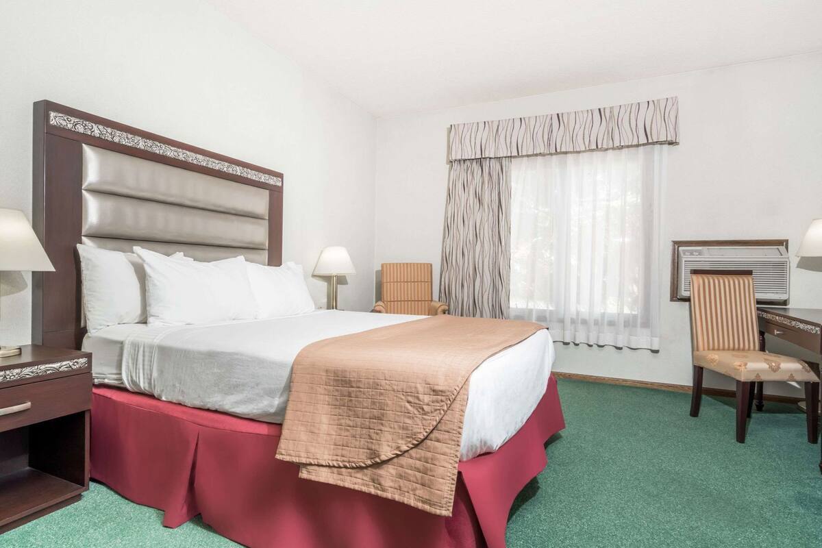 A hotel room with a bed, two bedside tables, a chair, a window with curtains, and a wall-mounted air conditioning unit.