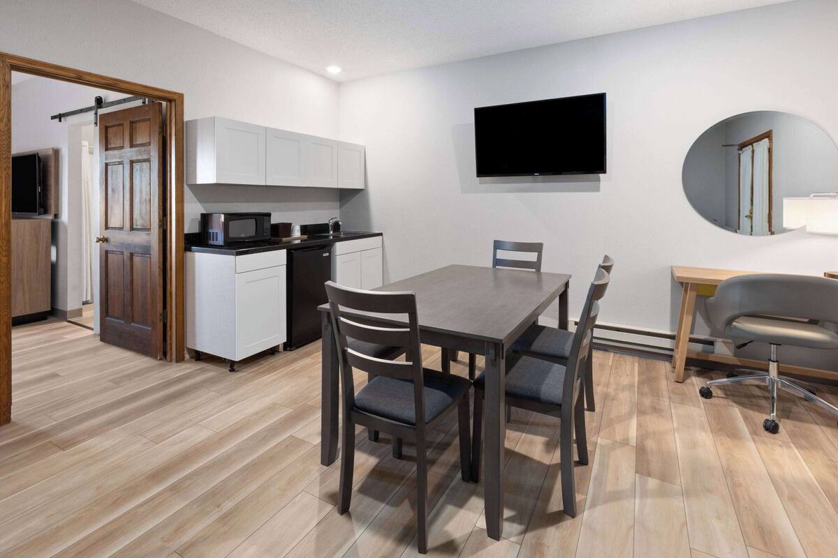 A modern kitchen with white cabinets, a black microwave, and a stove. There is a dining table with chairs and a TV mounted on the wall.