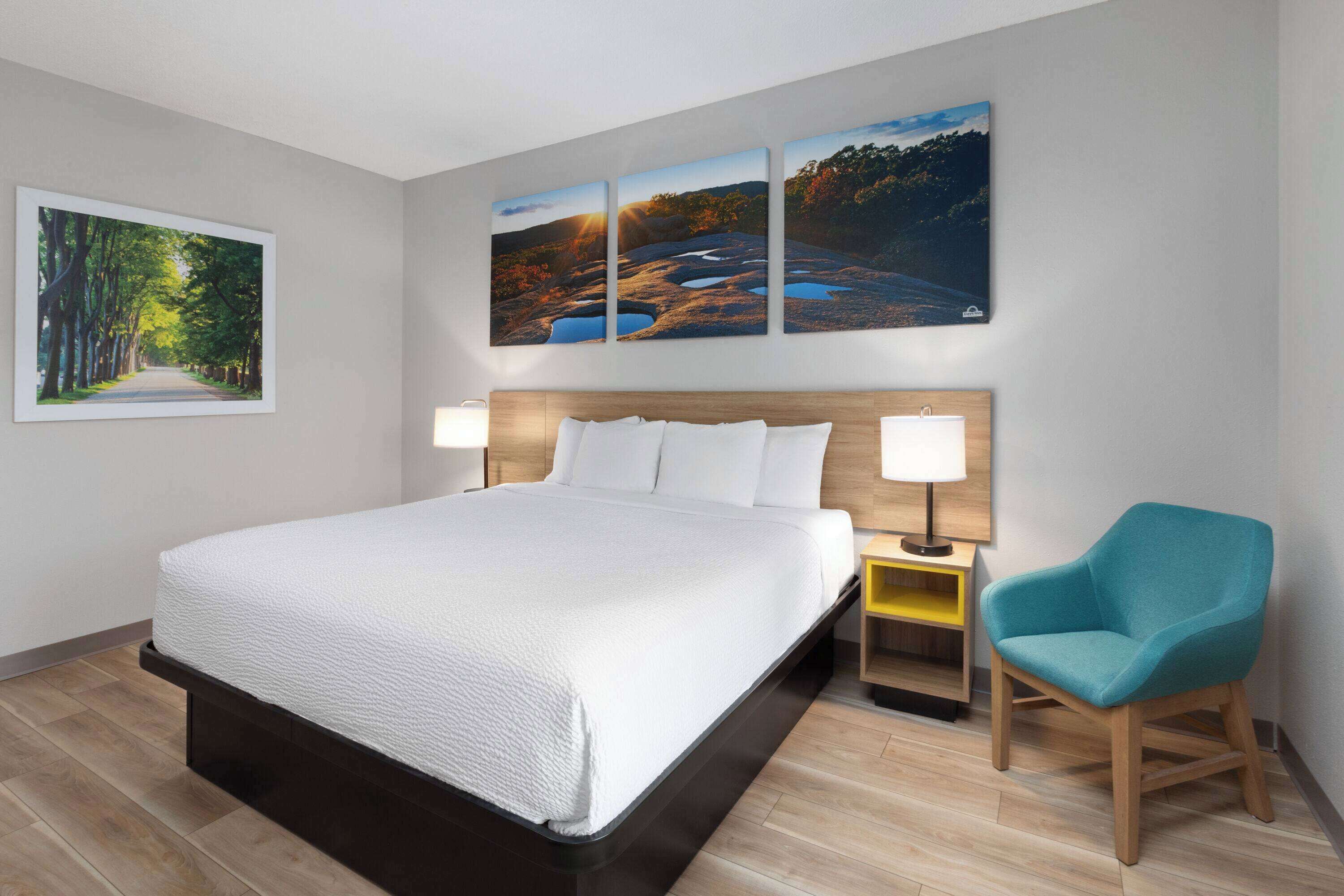 Une chambre d’hôtel avec un grand lit, deux tables de chevet, une chaise et trois tableaux représentant des paysages accrochés au mur.