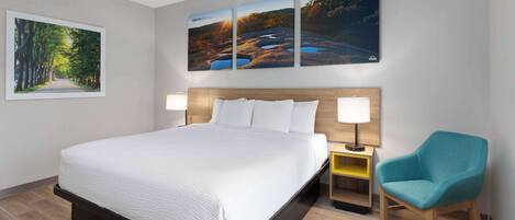 Une chambre d’hôtel avec un grand lit, deux tables de chevet, une chaise et trois tableaux représentant des paysages accrochés au mur.