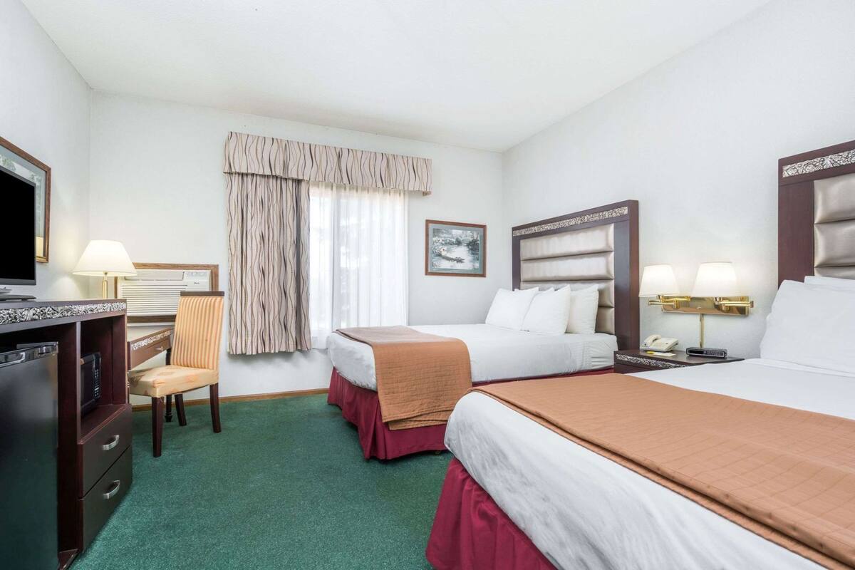 A hotel room with two beds, a desk with a television, a chair, a window with curtains, and a painting on the wall.