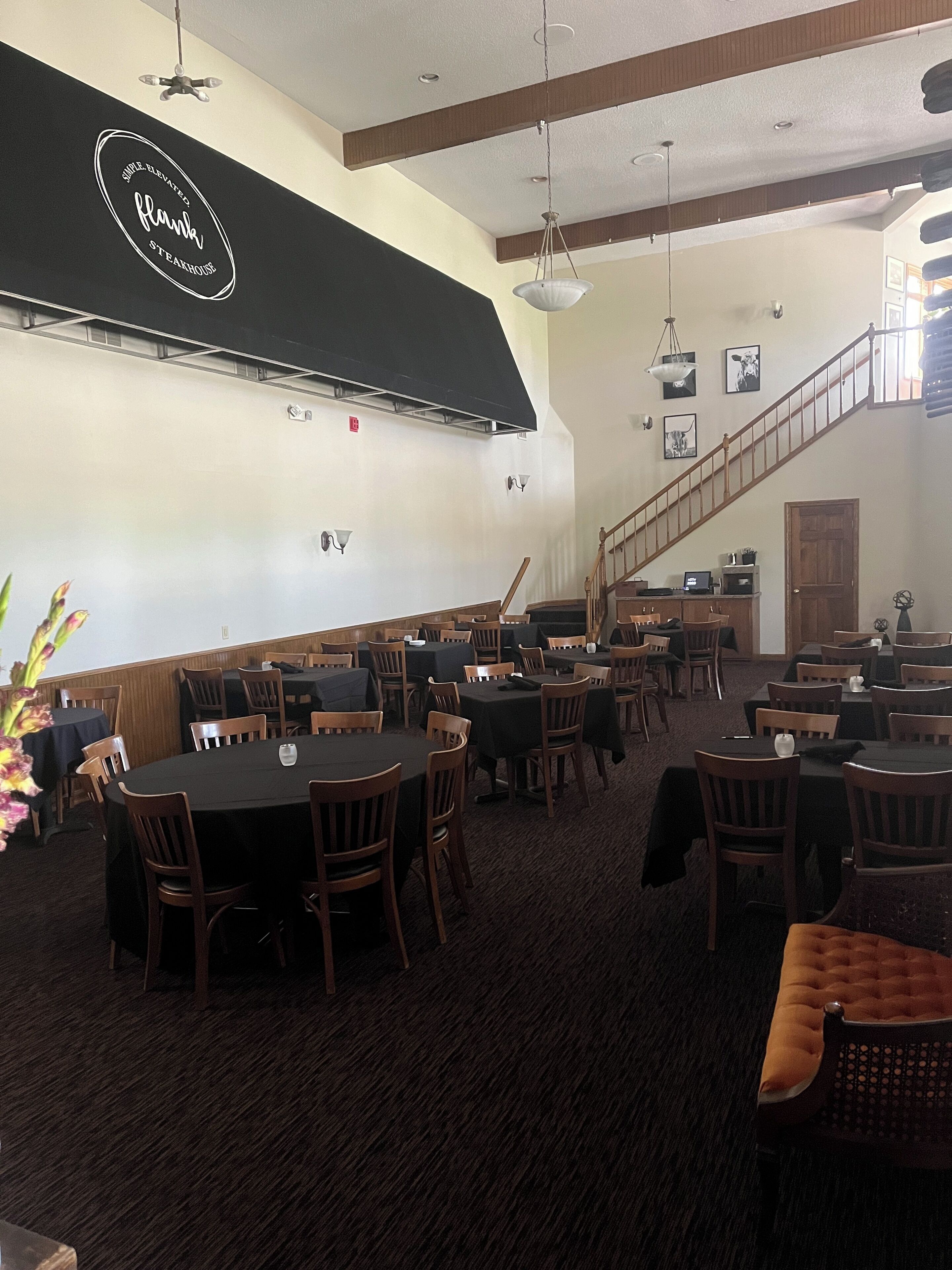A restaurant with round tables and wooden chairs, a staircase, and a black awning with the name "Flank".