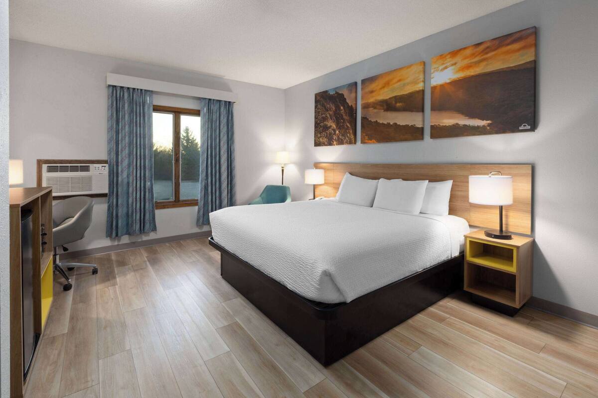 A hotel room with a bed, bedside tables, a desk, an air conditioning unit, and a window with curtains.