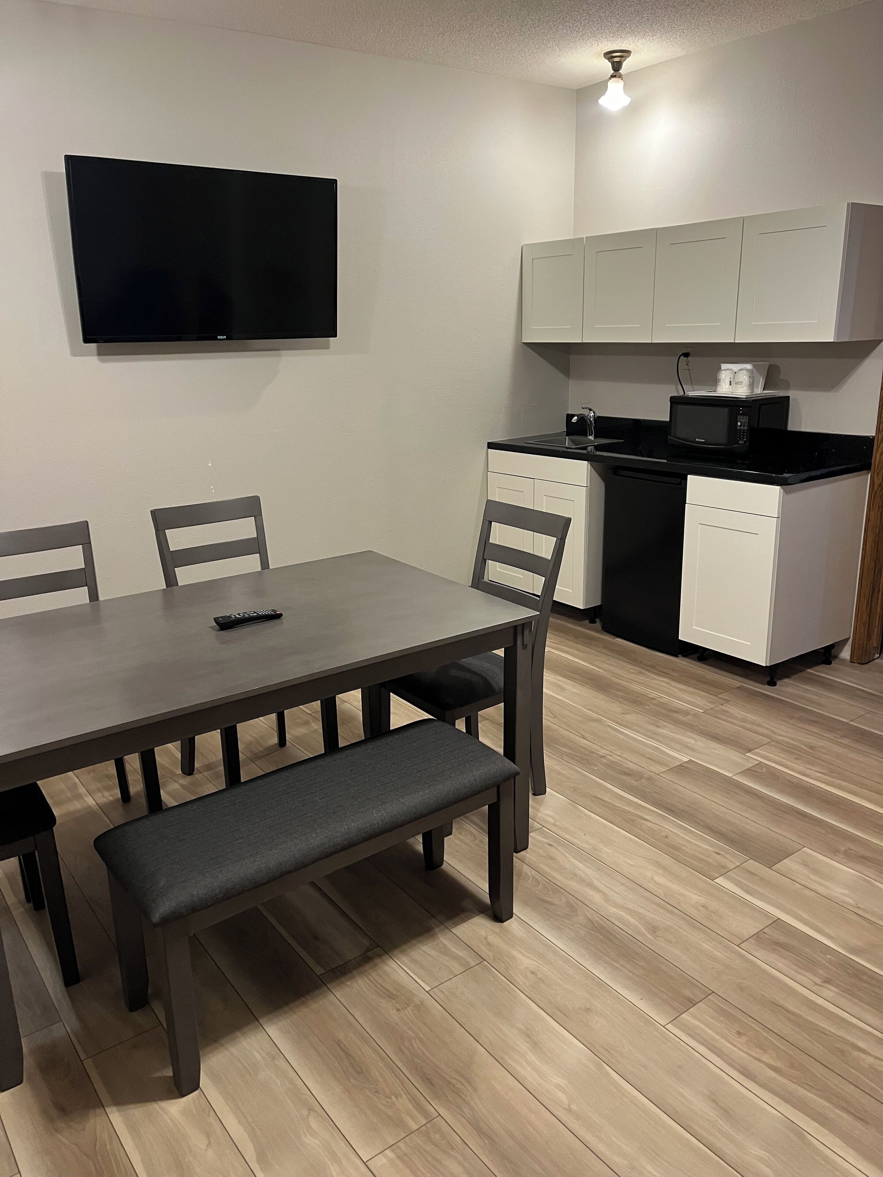 A compact living space with a dining table, chairs, a bench, a flat-screen TV, a microwave, and a small kitchen area with cabinets and a dishwasher.