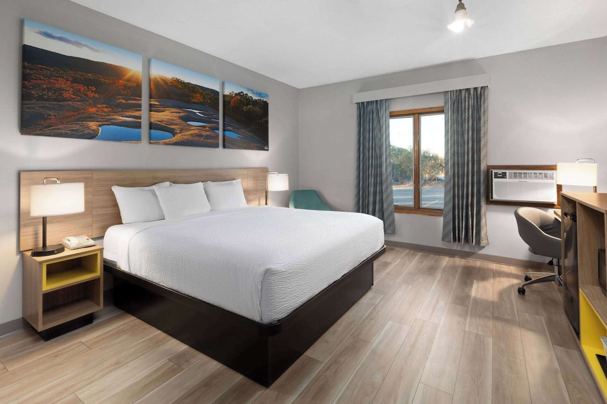 A hotel room with a large bed, bedside tables, a desk with a chair, a window with curtains, and wall art.