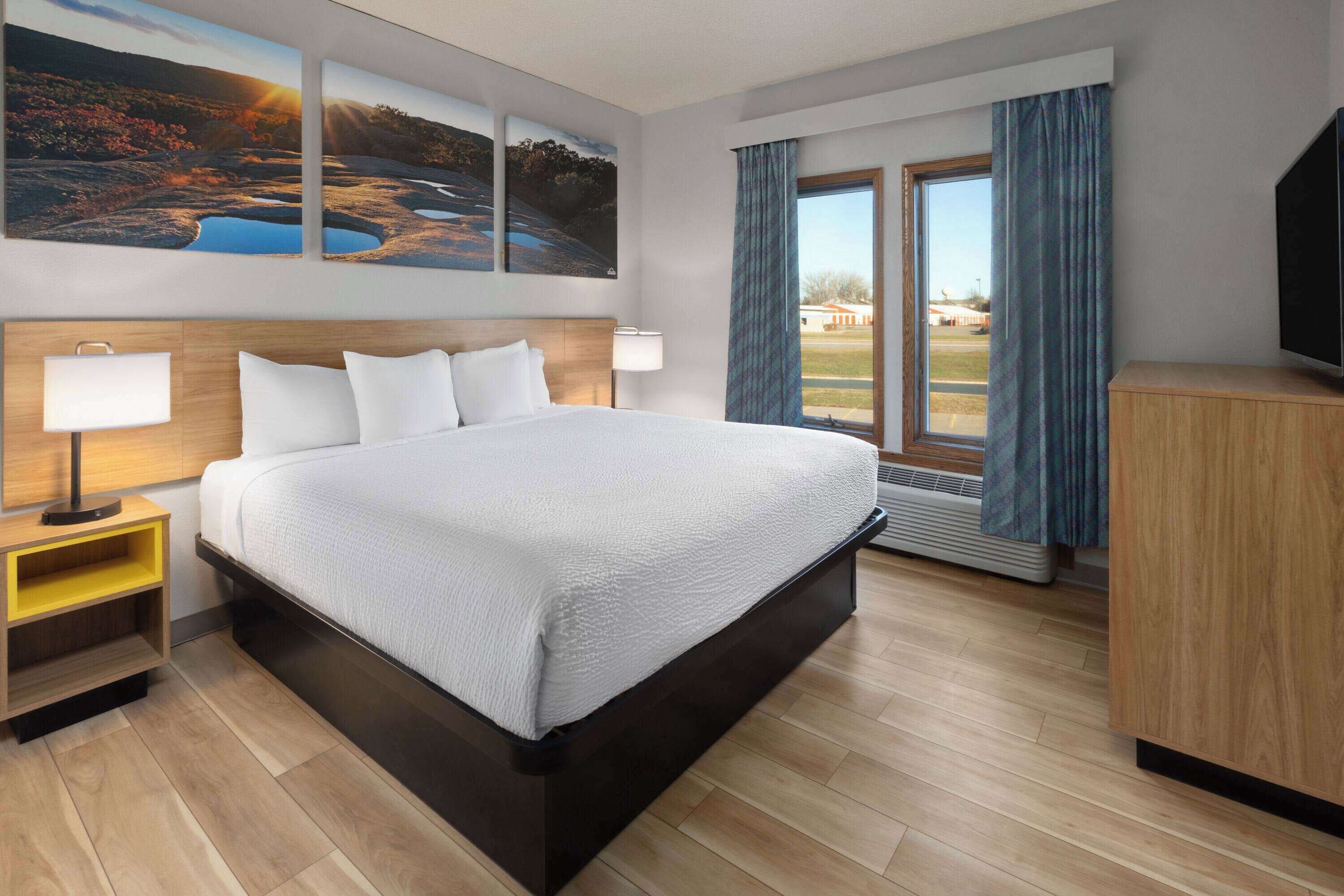 Hotelkamer met een groot bed, twee bedlampen, een houten nachtkastje, een flatscreen televisie en een groot raam met gordijnen.