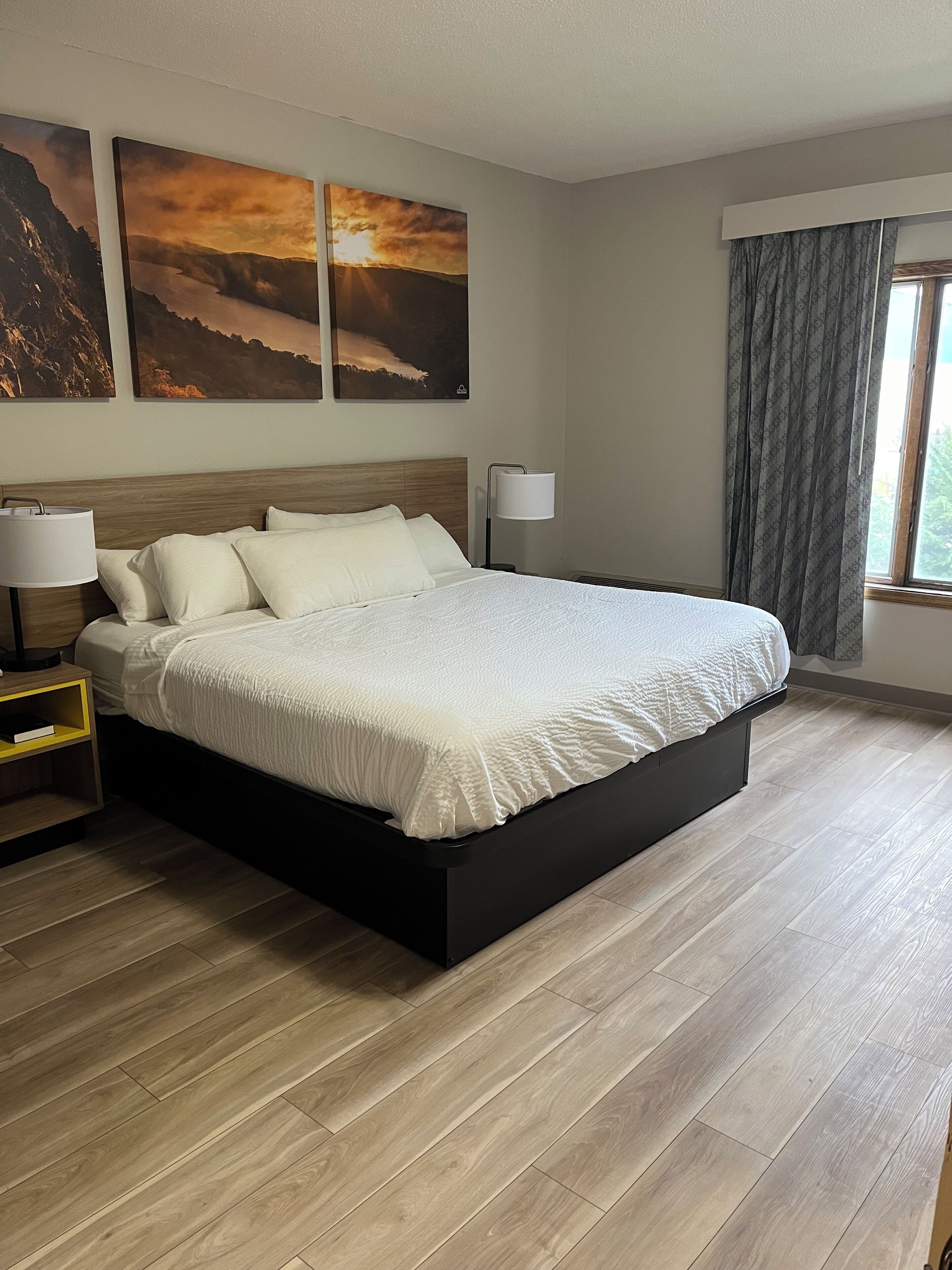 A modern hotel room with a large bed, two bedside tables, a window with curtains, and three landscape paintings on the wall.