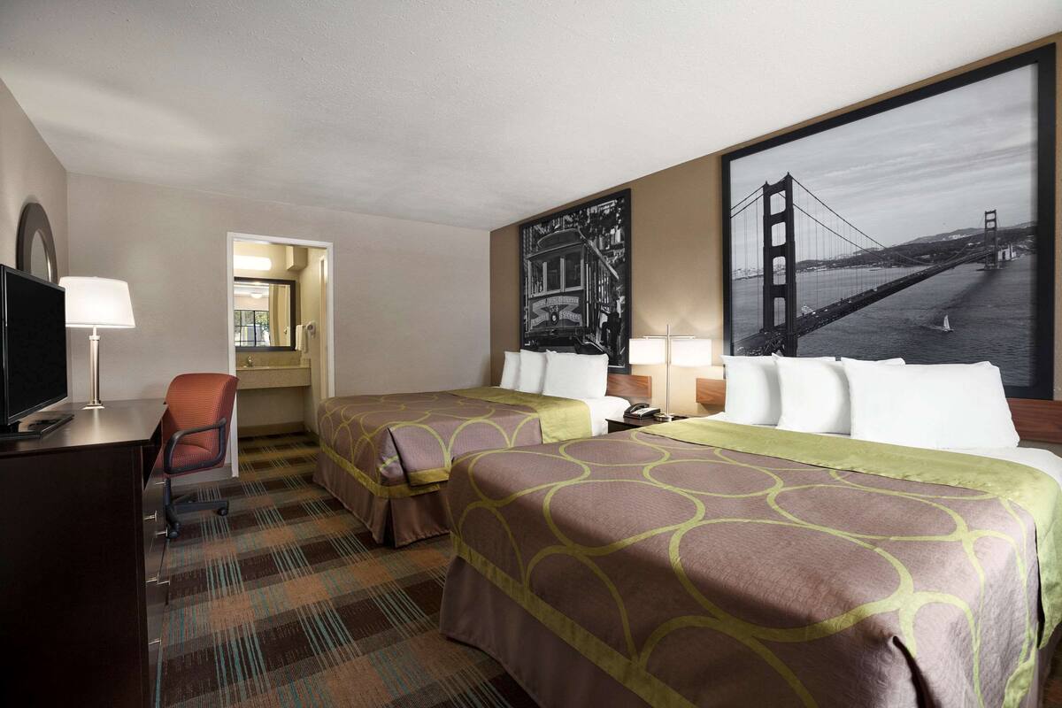 A hotel room with two beds, a desk, a chair, a TV, and a large wall mural of a bridge.