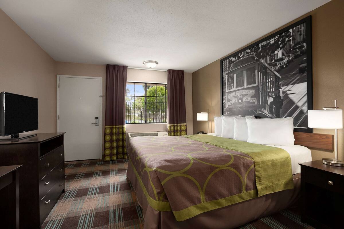 A hotel room with a bed, a television, a desk, a lamp, and a large framed picture on the wall.