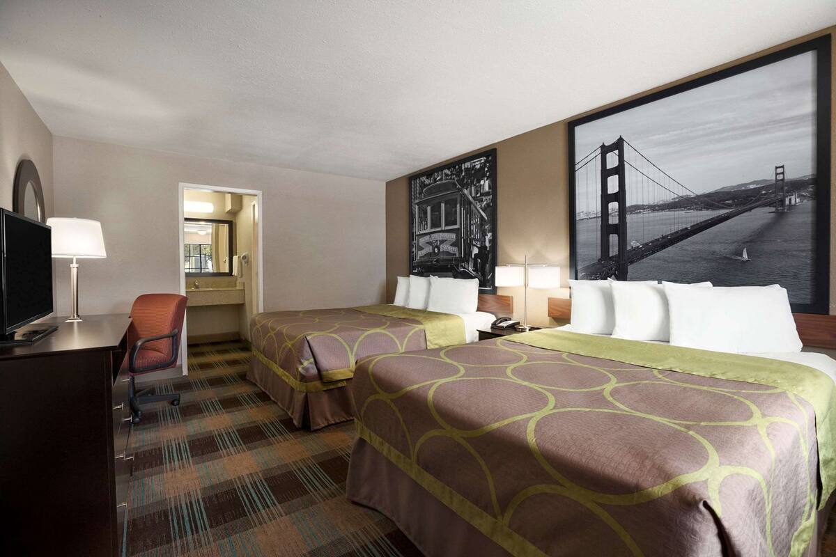 A hotel room with two beds, a desk with a TV, a chair, and a large window with a view of a bridge.