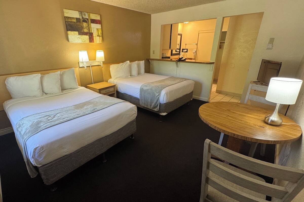 A hotel room with two beds, a small table with a lamp, a chair, a mirror, and a wall art piece.