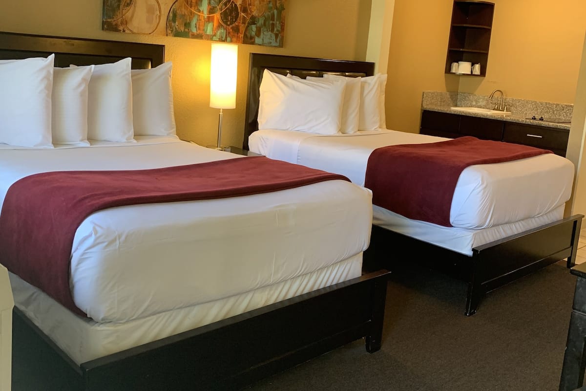A hotel room with two beds, a nightstand, a lamp, a painting, a shelf with toiletries, and a carpeted floor.