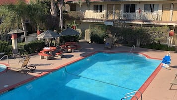 A swimming pool with lounge chairs and umbrellas, surrounded by palm trees and a building with a red roof.