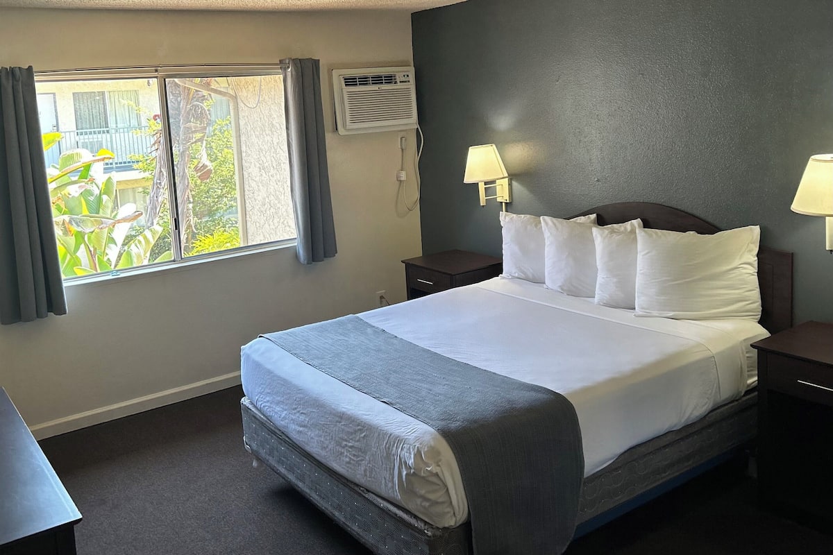 A hotel room with a bed, two bedside tables, a desk, a window with curtains, an air conditioning unit, and two wall-mounted lamps.