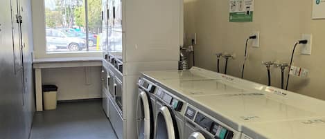 A laundry facility with multiple washing machines and dryers, a window with a view of parked cars, and a poster on the wall.