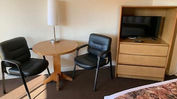 A hotel room with two black leather chairs, a wooden table, a white lamp, a television on a wooden cabinet, and a bed with a patterned bedspread.
