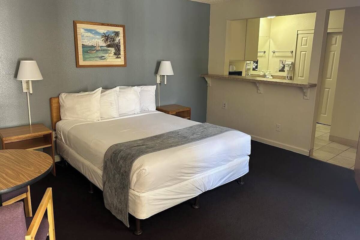 A hotel room with a bed, two bedside tables, a round table, a chair, a painting, a mirror, and a bathroom visible through an open door.