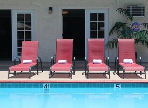 Zona de piscina con cuatro sillas reclinables rojas, cada una con una toalla blanca, y un letrero en la pared que dice 'MANTENER CERRADO'.