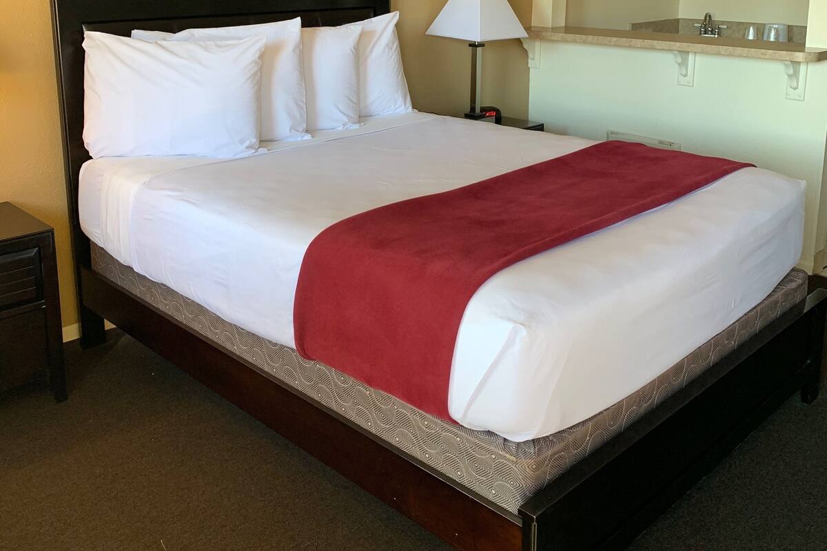 A hotel room with a large bed, a nightstand, a lamp, a painting on the wall, and a bathroom with a mirror and sink.